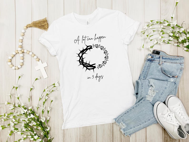May include: White t-shirt featuring the text "A lot can happen in 3 days" and a black graphic of a crown of thorns and a wreath. The shirt is styled with blue jeans and white trainers.