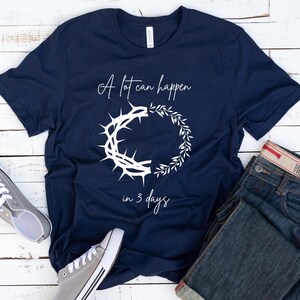 May include: Navy blue t-shirt with white text and graphic. The text reads "A lot can happen in 3 days" with a crown of thorns and a wreath design. Grey canvas trainers and blue jeans are also in the image.
