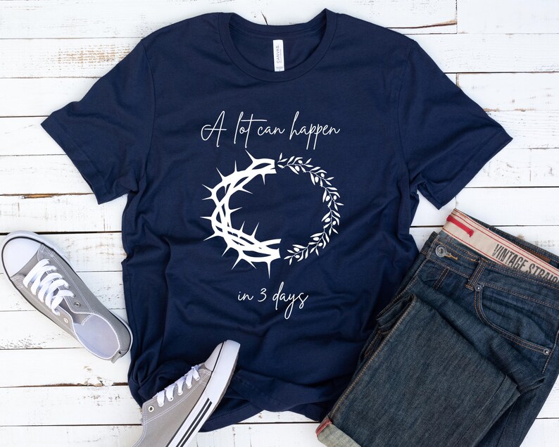 May include: Navy blue t-shirt with white text and graphic. The text reads "A lot can happen in 3 days" with a crown of thorns and a wreath design. Grey canvas trainers and blue jeans are also in the image.
