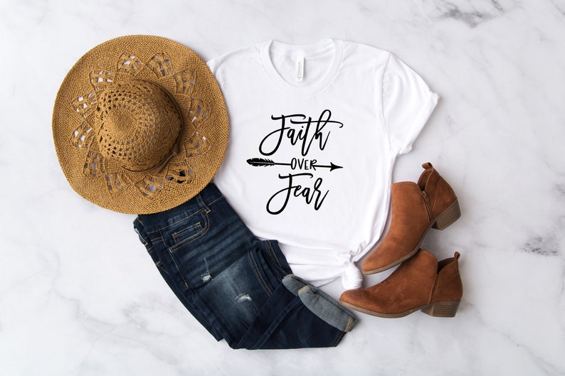 May include: White t-shirt with the text "Faith over Fear" in black script with an arrow between the words. The shirt is styled with blue jeans, a brown hat, and brown ankle boots.