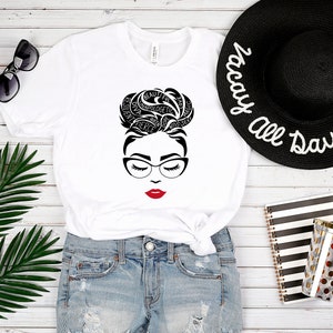 May include: A white t-shirt featuring a black graphic of a face with glasses and a messy bun hairstyle composed of words including beautiful, resilient, diva, unapologetic, fierce, queen, dope, boss, sexy, love, and magic, with bright red lips.