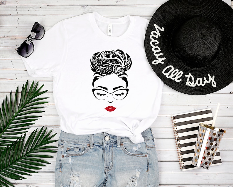 May include: A white t-shirt featuring a black graphic of a face with glasses and a messy bun hairstyle composed of words including beautiful, resilient, diva, unapologetic, fierce, queen, dope, boss, sexy, love, and magic, with bright red lips.
