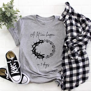 May include: A grey t-shirt with the text "A lot can happen in 3 days" and a graphic of a crown of thorns and a wreath. Black and white Converse trainers and a black and white checked shirt are also in the image.