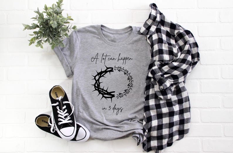 May include: A grey t-shirt with the text "A lot can happen in 3 days" and a graphic of a crown of thorns and a wreath. Black and white Converse trainers and a black and white checked shirt are also in the image.