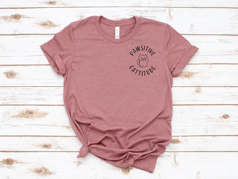 Puede incluir: Una camiseta de color malva con un gr&aacute;fico negro de un gato con el texto "Pawsitive Cattitude" en la parte delantera.