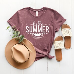 May include: A mauve t-shirt with the text "hello summer" in white lettering. The shirt is styled with a brown hat and white sandals.