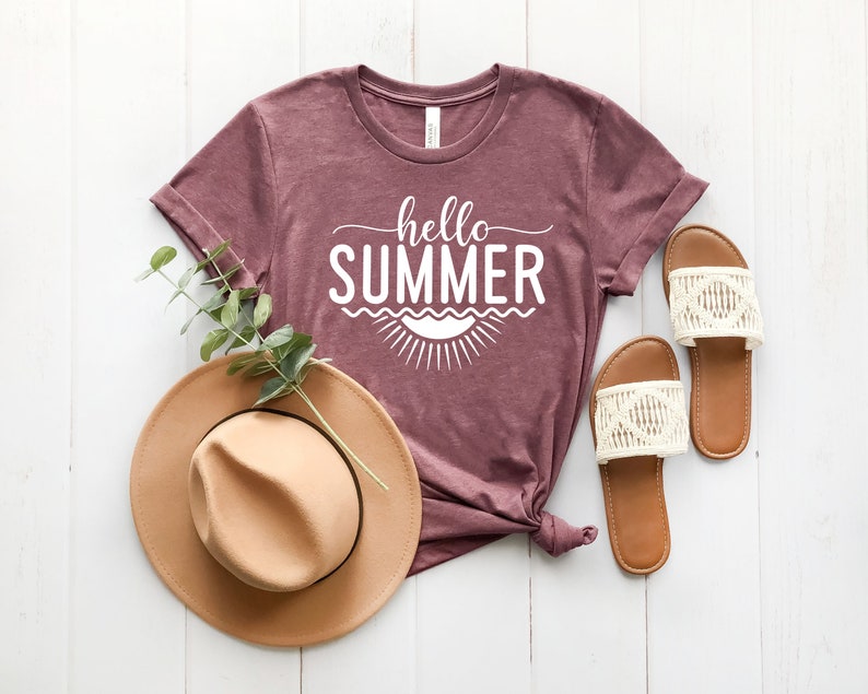 May include: A mauve t-shirt with the text "hello summer" in white lettering. The shirt is styled with a brown hat and white sandals.