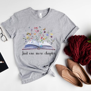 Op de afbeelding: Grijs T-shirt met een aquarel-illustratie van een open boek met bloemen en vlinders die eruit groeien. De tekst "Just one more chapter" is onder het boek gedrukt.