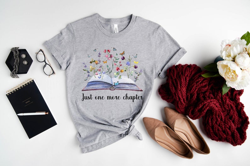 Op de afbeelding: Grijs T-shirt met een aquarel-illustratie van een open boek met bloemen en vlinders die eruit groeien. De tekst "Just one more chapter" is onder het boek gedrukt.
