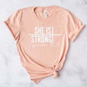 May include: A peach-colored t-shirt with the text "SHE IS STRONG" and "proverbs 31:25" printed in white.