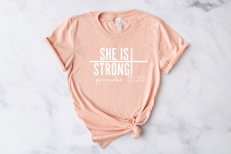 May include: A peach-colored t-shirt with the text "SHE IS STRONG" and "proverbs 31:25" printed in white.