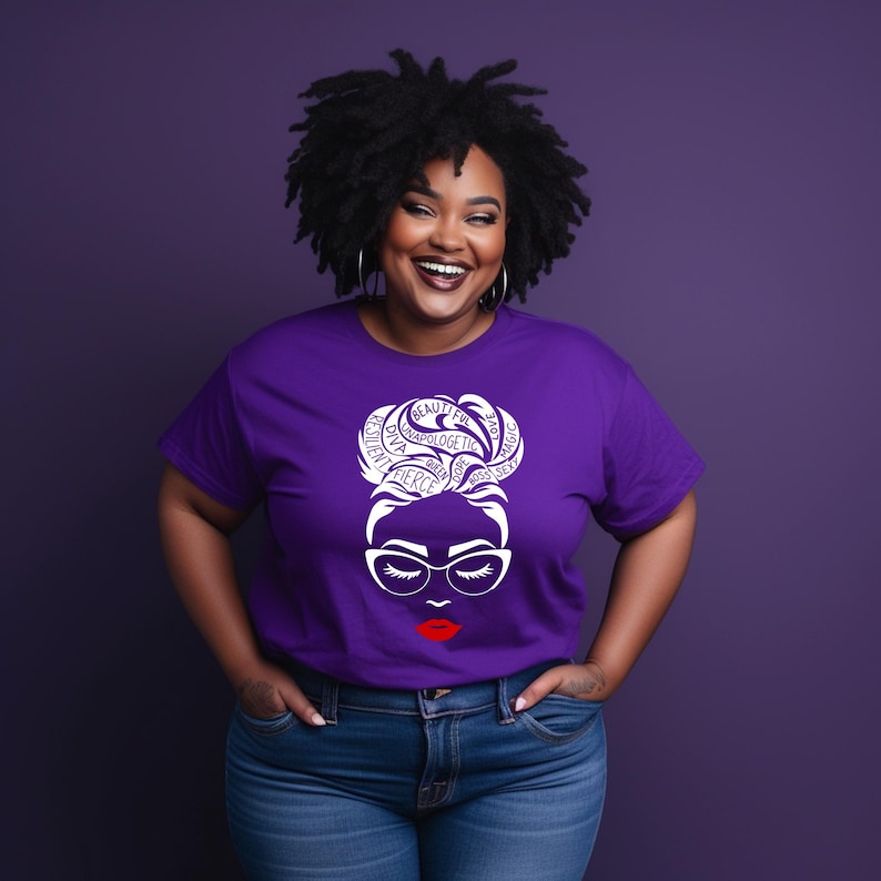 May include: A purple short-sleeved t-shirt featuring a white graphic of a face with glasses and a high bun hairstyle. The hair design incorporates the words resilient, diva, unapologetic, queen, fierce, dope, boss, sexy, beautiful, love, and magic.