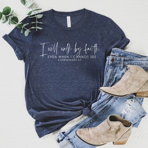May include: Navy blue t-shirt with the text "I will walk by faith, even when I cannot see, 2 Corinthians 5:7" in white script. The shirt is paired with blue jeans and tan ankle boots.