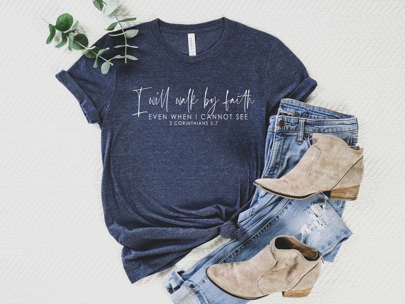 May include: Navy blue t-shirt with the text "I will walk by faith, even when I cannot see, 2 Corinthians 5:7" in white script. The shirt is paired with blue jeans and tan ankle boots.
