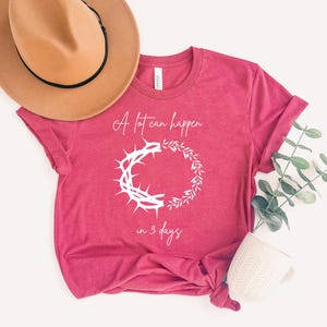May include: A magenta t-shirt with the text "A lot can happen in 3 days." The shirt displays a white graphic of a crown of thorns and a wreath. A tan hat with a black band and a white mug are also visible.