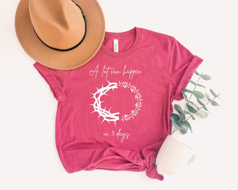 May include: A magenta t-shirt with the text "A lot can happen in 3 days." The shirt displays a white graphic of a crown of thorns and a wreath. A tan hat with a black band and a white mug are also visible.