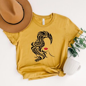 May include: A mustard yellow t-shirt with a black silhouette of a woman's head with the words "fuerte, dama, poderosa, latina, mujer, trabajadora, capa" surrounding her hair.