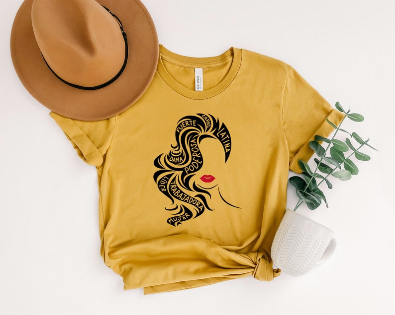 May include: A mustard yellow t-shirt with a black silhouette of a woman's head with the words "fuerte, dama, poderosa, latina, mujer, trabajadora, capa" surrounding her hair.