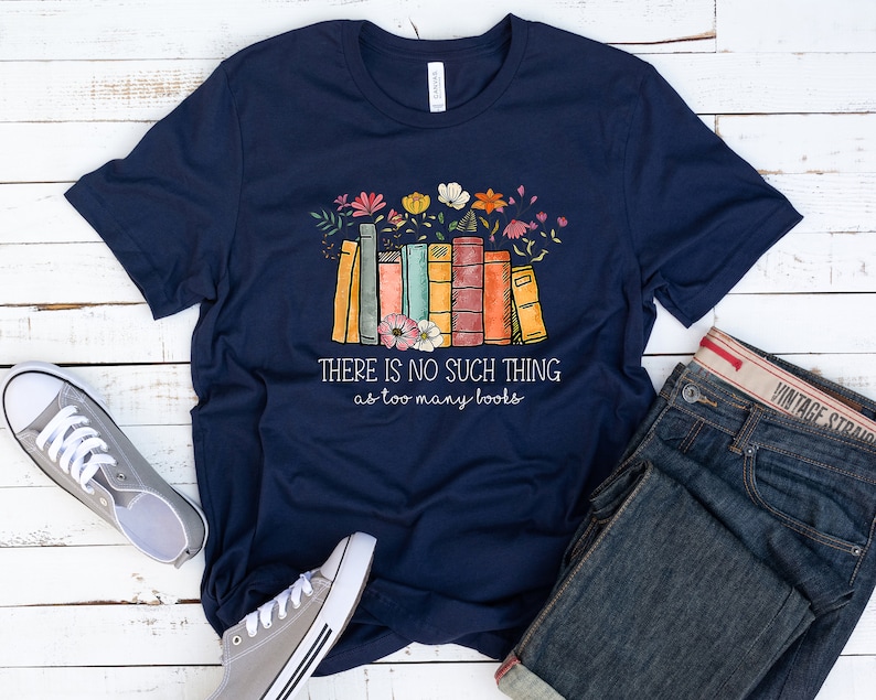 May include: Navy blue t-shirt with a stack of colorful books and flowers. The text on the shirt reads "There is no such thing as too many books."
