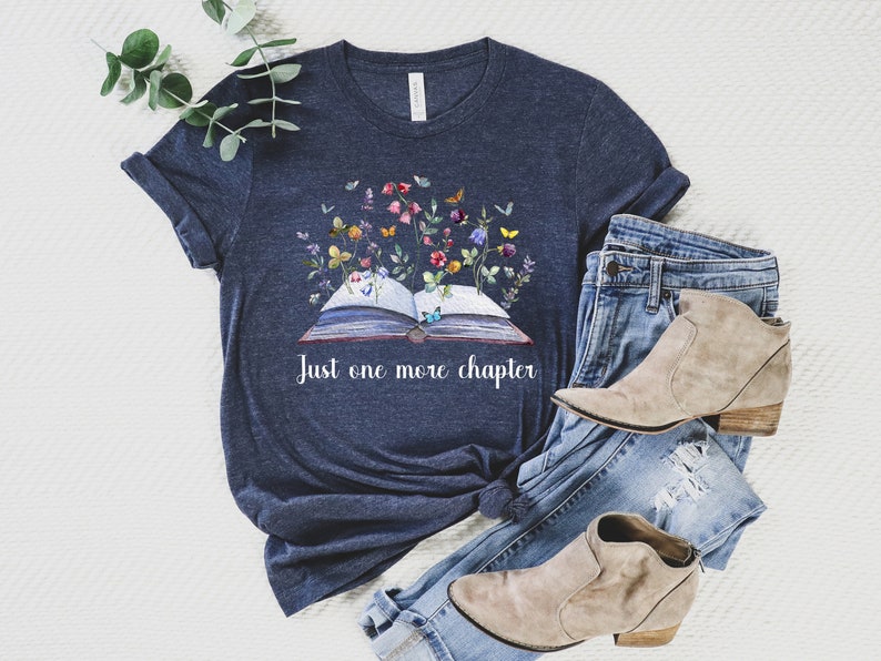 Op de afbeelding: Een marineblauw t-shirt met een afbeelding van een open boek met bloemen en vlinders die eruit groeien. De tekst "Just one more chapter" is onder het boek gedrukt. Het shirt ligt op een wit oppervlak met een paar lichtbruine enkellaarsjes en een blauwe jeans.