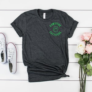 Puede incluir: Una camiseta gris oscuro jaspeada con un gr&aacute;fico verde de un gato con el texto "Pawsitive Cattitude".