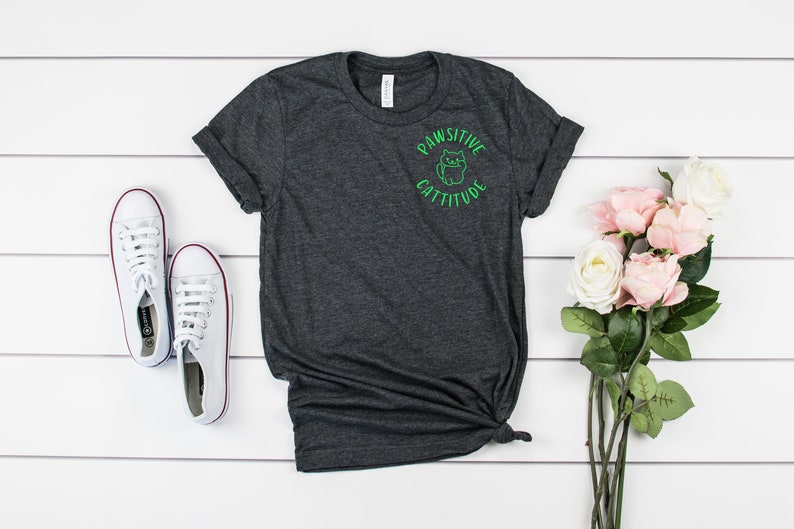 Puede incluir: Una camiseta gris oscuro jaspeada con un gr&aacute;fico verde de un gato con el texto "Pawsitive Cattitude".