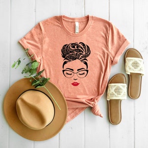 May include: A peach-colored t-shirt featuring a black graphic of a face with glasses and a messy bun hairstyle formed by words including beautiful, love, magic, fierce, and queen. The lips are red. A tan hat and two beige sandals are nearby.