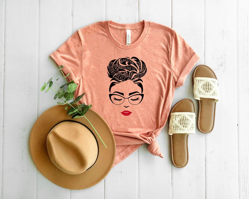 May include: A peach-colored t-shirt featuring a black graphic of a face with glasses and a messy bun hairstyle formed by words including beautiful, love, magic, fierce, and queen. The lips are red. A tan hat and two beige sandals are nearby.