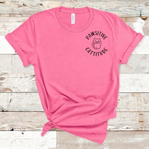Puede incluir: Una camiseta rosa con un gr&aacute;fico negro que dice "Pawsitive Cattitude" con un gato de dibujos animados dentro del c&iacute;rculo.