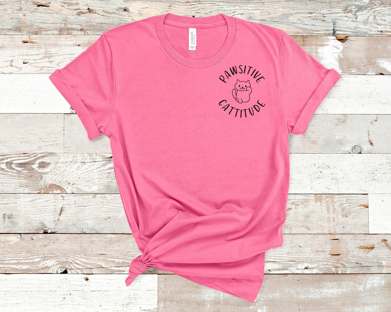 Puede incluir: Una camiseta rosa con un gr&aacute;fico negro que dice "Pawsitive Cattitude" con un gato de dibujos animados dentro del c&iacute;rculo.