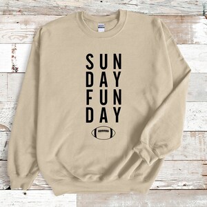 Pu&ograve; includere: Felpa beige con testo nero che recita "SUN DAY FUN DAY" e un grafico di un pallone da calcio nero sotto il testo.