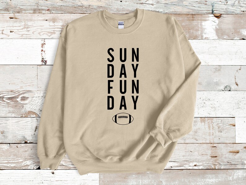 Pu&ograve; includere: Felpa beige con testo nero che recita "SUN DAY FUN DAY" e un grafico di un pallone da calcio nero sotto il testo.