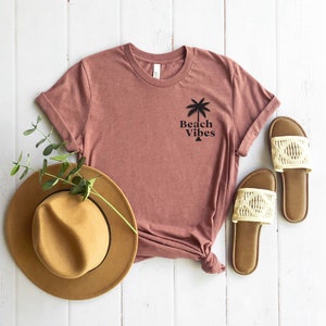 May include: A mauve t-shirt with a palm tree graphic and the text "Beach Vibes". The shirt is laid out on a white wooden surface with a brown hat and white sandals.