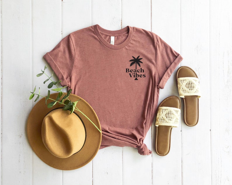 May include: A mauve t-shirt with a palm tree graphic and the text "Beach Vibes". The shirt is laid out on a white wooden surface with a brown hat and white sandals.