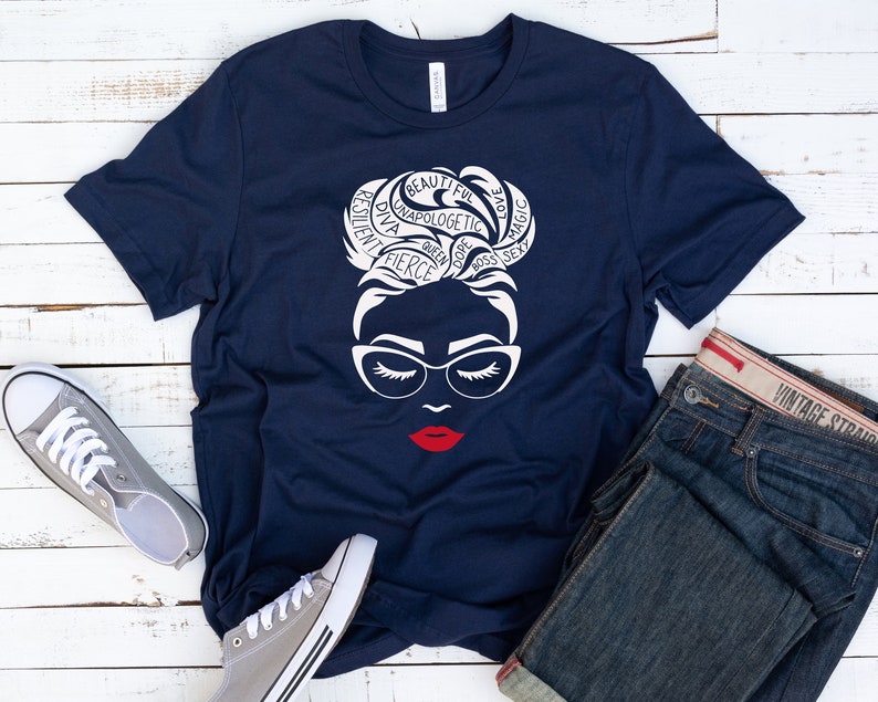 May include: A navy blue t-shirt featuring a white graphic of a person with a high bun hairstyle, cat-eye glasses, and bright red lips. The hair design incorporates the words beautiful, unapologetic, love, magic, resilient, diva, queen, dope, boss, and sexy.