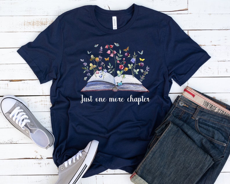 Op de afbeelding: Een marineblauw t-shirt met een witte afbeelding van een open boek met bloemen en vlinders die eruit groeien. De tekst "Just one more chapter" staat onder het boek.