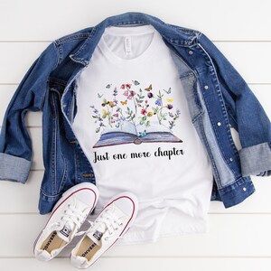 Op de afbeelding: Wit T-shirt met een afbeelding van een open boek met bloemen en vlinders die eruit groeien. De tekst "Just one more chapter" staat onder het boek.