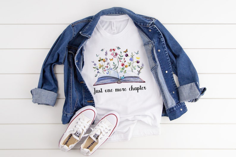 Op de afbeelding: Wit T-shirt met een afbeelding van een open boek met bloemen en vlinders die eruit groeien. De tekst "Just one more chapter" staat onder het boek.