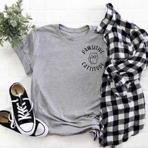 Puede incluir: Camiseta gris con un gr&aacute;fico negro de un gato y el texto "Pawsitive Cattitude". La camiseta se combina con una camisa de cuadros blanco y negro y zapatillas negras.
