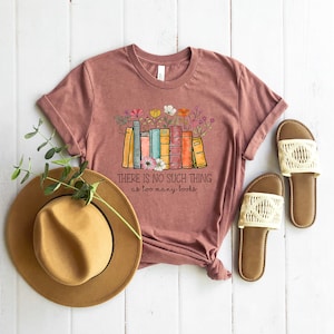 May include: A maroon t-shirt with a graphic of a stack of colorful books with flowers. The text on the shirt reads "There is no such thing as too many books".