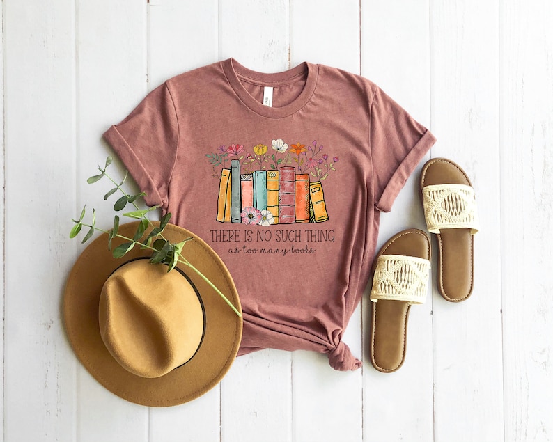 May include: A maroon t-shirt with a graphic of a stack of colorful books with flowers. The text on the shirt reads "There is no such thing as too many books".