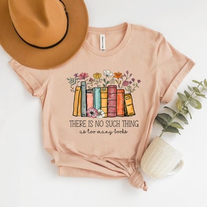 May include: A light pink t-shirt with a graphic of a stack of colorful books with flowers on top. The text "THERE IS NO SUCH THING as too many books" is printed below the graphic.