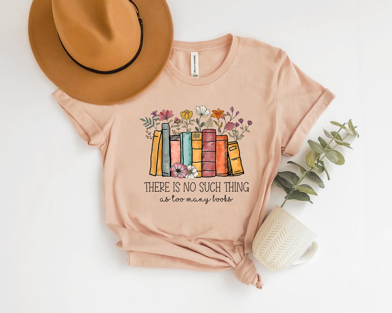May include: A light pink t-shirt with a graphic of a stack of colorful books with flowers on top. The text "THERE IS NO SUCH THING as too many books" is printed below the graphic.