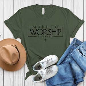 Puede incluir: Una camiseta verde con el texto "MADE TO WORSHIP - PSALMS 95:1" en letras negras. La camiseta est&aacute; colocada sobre una superficie de madera blanca con un sombrero marr&oacute;n, unos vaqueros azules y unas zapatillas blancas.