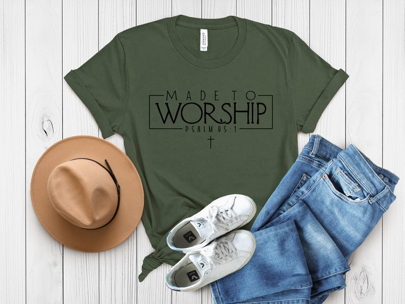 Puede incluir: Una camiseta verde con el texto "MADE TO WORSHIP - PSALMS 95:1" en letras negras. La camiseta est&aacute; colocada sobre una superficie de madera blanca con un sombrero marr&oacute;n, unos vaqueros azules y unas zapatillas blancas.