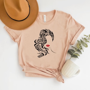 May include: A light pink t-shirt with a black silhouette of a woman's face with red lips. The woman's hair is styled in a way that forms the words "fuerte", "rosa", "poderosa", "dama", "trabajadora", "mujer", "capaz", and "latina" in Spanish.