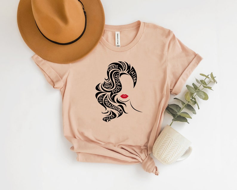 May include: A light pink t-shirt with a black silhouette of a woman's face with red lips. The woman's hair is styled in a way that forms the words "fuerte", "rosa", "poderosa", "dama", "trabajadora", "mujer", "capaz", and "latina" in Spanish.