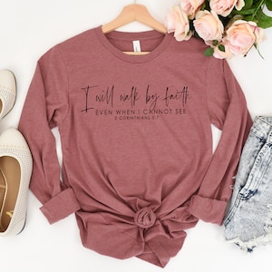 May include: A long-sleeved, mauve-colored shirt with the text "I will walk by faith EVEN WHEN I CANNOT SEE 2 CORINTHIANS 5:7" in black script. The shirt is displayed with a pair of cream-colored flats and denim shorts.