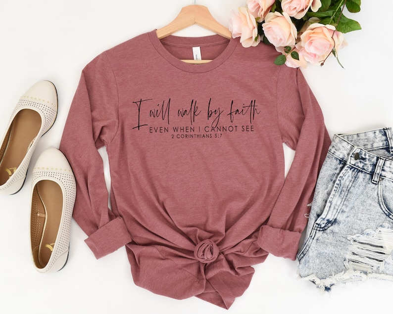 May include: A long-sleeved, mauve-colored shirt with the text "I will walk by faith EVEN WHEN I CANNOT SEE 2 CORINTHIANS 5:7" in black script. The shirt is displayed with a pair of cream-colored flats and denim shorts.