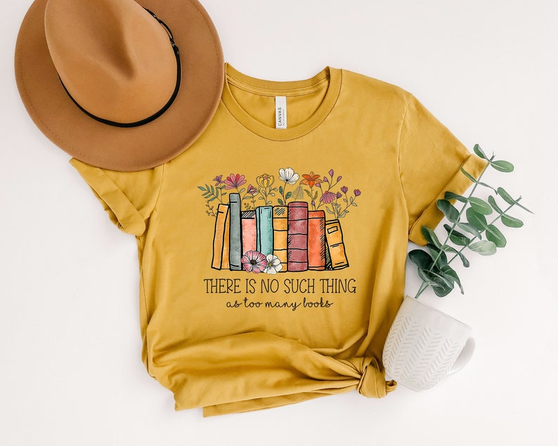 May include: A mustard yellow t-shirt with a graphic of a stack of books with flowers on top. The text "There is no such thing as too many books" is printed below the graphic.